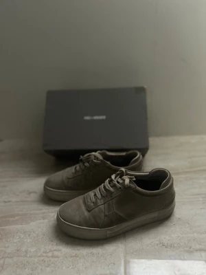 Axel arigato - Snygga beigea sneakers i mocka med klassisk låg profil och matchande snörning. Skorna har en enkel design med diskreta sömmar och vit sula. Perfekta för dig som gillar stilrena och neutrala färger.