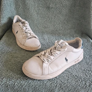 Vita Polo Ralph Lauren sneakers - Snygga vita sneakers från Polo Ralph Lauren med klassisk låg profil. Skorna har vita snören med Polo-logga och den ikoniska hästloggan på sidan. Tillverkade i slätt läder med vit sula. Perfekta för en clean och stilren look.