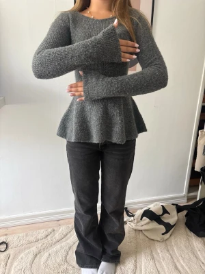 Grå peplum stickad tröja - Mysig grå stickad tröja med peplumskärning och långa ärmar. Tröjan har rund halsringning och är figurnära upptill med en utsvängd nederdel. Perfekt för dig som gillar en unik och trendig siluett.