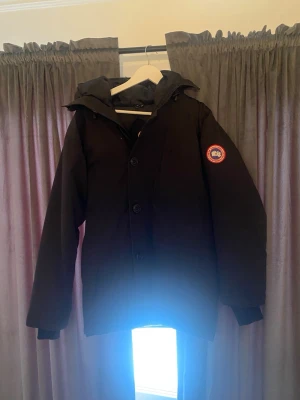Svart dunjacka med huva Canada Goose - Säljer en äkta svart dunjacka från Canada Goose med avtagbar huva och klassisk logotyp på ärmen. Jackan har dragkedja och knappar framtill samt ribbade muddar vid ärmsluten. Perfekt för kalla vinterdagar och riktigt varm tack vare dunfyllningen. Priset är förhandlingsbart!