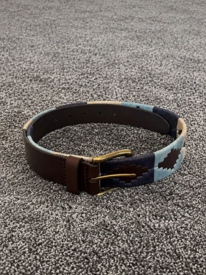 Brunt och blått mönstrat bälte - Snyggt bälte i brunt skinn med blått och beige geometriskt mönster. Bältet har en metallspänne i guldton och detaljer i olika blå nyanser. Perfekt för att lyfta din outfit med en cool accessoar.