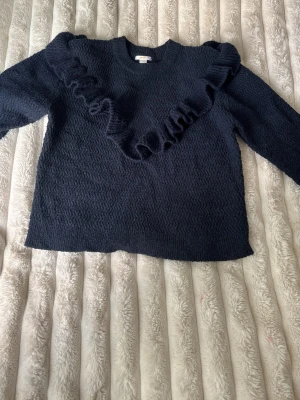 Mörkblå stickad tröja med volang - Snygg mörkblå stickad tröja med vågig struktur och en stor volangdetalj som går diagonalt över framsidan och axlarna. Tröjan har rund halsringning och långa ärmar. Perfekt för dig som vill ha något extra i garderoben.