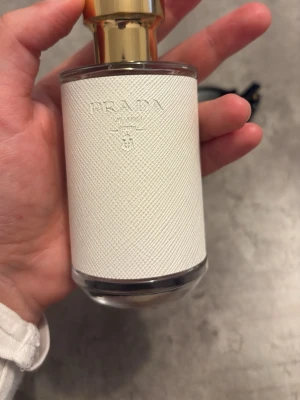 Prada parfym med vit textur - Elegant parfymflaska från Prada med vit texturerad yta och guldfärgat lock. Flaskan är genomskinlig med en exklusiv känsla och har Pradas logga diskret präglad på framsidan. Perfekt för dig som gillar lyxiga dofter och snygg design.