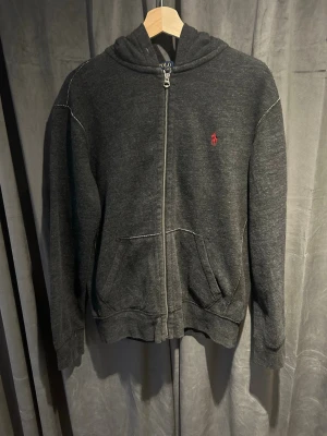 Mörkgrå Ralph Lauren zip hoodie - Mörkgrå hoodie med dragkedja och huva. Har en liten röd broderad logga på bröstet och vita kontrastsömmar. Två fickor framtill och ribbade muddar vid ärmar och nederkant. Perfekt för en avslappnad och stilren look.