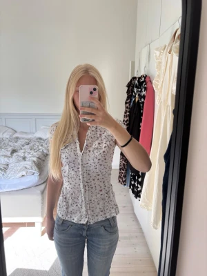 Vit blommig blus - Söt vit blus med små blommor i rosa och lila toner. Blusen har knappar framtill, volangkant längs knäppningen och är lätt figursydd. Man kan knyta den baktill.