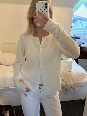 Vit basic kofta med rund hals - En stilren vit kofta med rund halsringning och knäppning framtill. Koftan har långa ärmar och en mjuk, stickad känsla. Perfekt att matcha med jeans eller kjol för en clean look.