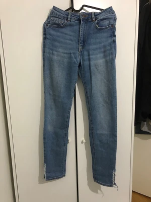 Ljusblå skinny jeans med råa kanter - Säljer ett par ljusblå skinny jeans med hög midja och slitna, råa kanter vid bensluten. Jeansen har klassisk femficksdesign och är tillverkade i stretchigt denimtyg för en bekväm passform. Perfekta för en avslappnad och trendig look.