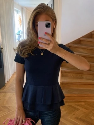 Mörkblå peplum topp med volang - Snygg mörkblå topp med peplum och dubbla volanger nedtill. Kortärmad och figurnära passform som ger en feminin siluett. Perfekt att styla med jeans eller kjol för en trendig look.