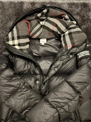 Svart dunväst från Burberry - Snygg svart dunväst från Burberry med klassiskt rutigt foder i grått, vitt, svart och rött. Västen har huva, dragkedja och tryckknappar samt logga på bröstet. Perfekt för lager på lager och streetstyle.