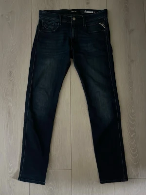 Mörkblå jeans från Replay - Snygga mörkblå  jeans från Replay i storlek 32/32 med klassisk femficksdesign och subtila slitningar framtill. Jeansen har dragkedjegylf, silverfärgade detaljer och en läderpatch med logga bak i midjan. Tillverkade i stretchig denim för extra komfort. Sparsamt använda