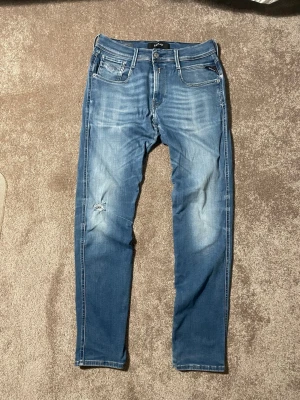 Blå slim fit jeans från Replay - Säljer ett par blå slim fit jeans från Replay med slitningar och snygg tvätt. Jeansen har fem fickor, dragkedjegylf och en liten reva på vänster ben för en trendig look. Perfekta för dig som gillar en avslappnad men stilren stil.