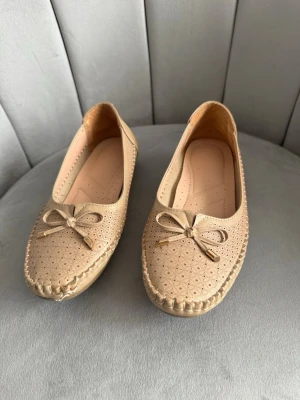 Beige ballerinaskor med rosett - Snygga beige ballerinaskor med dekorativ rosett framtill och perforerat mönster över hela skon. Skorna har rundad tå och sömmar längs kanten för en klassisk look. Tillverkade i ett mjukt material som ser ut som konstläder.