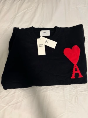 Svart stickad tröja från Ami Paris  - Snygg svart stickad tröja från Ami Paris med röd hjärt- och A-detalj på bröstet. Klassisk rund halsringning och ribbade muddar. Perfekt för dig som vill ha en stilren men ändå unik look.