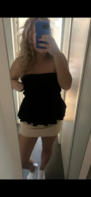 Svart axelbandslös volangtop - Säljer en svart axelbandslös topp med volangkant och smockad överdel. Toppen har en bandeauformad passning och är perfekt för dig som vill ha en trendig och stilren look. Materialet känns lätt och luftigt, vilket gör den skön att bära under varma dagar.