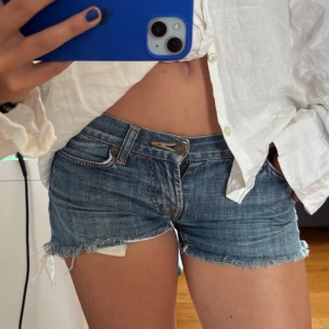 Blå jeansshorts med fransig kant - Säljer ett par klassiska blå jeansshorts med rå, fransig kant och femficksmodell. Shortsen har låg midja och slitna detaljer för en avslappnad look. Perfekta till sommaren och lätta att matcha med olika toppar.