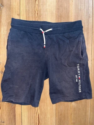 Mörkblå shorts från Tommy Hilfiger - Mörkblå shorts från Tommy Hilfiger med vit snörning i midjan och tryckt logga på vänster ben. Tillverkade i mjuk bomull, har sidofickor och en bakficka. Perfekta för sommardagar och avslappnade tillfällen. Lite solblekta och använda under en sommar därav anpassar jag priset utefter det självfallet! Nypris:899kr.  Mitt pris:299