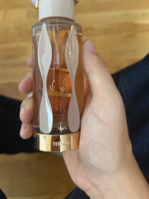 Lyxig parfymflaska med gulddetaljer - Snygg parfymflaska i genomskinligt glas med guld- och vita detaljer. Flaskan har en elegant form och ett dekorativt lock. Innehållet är bärnstensfärgat och designen känns exklusiv och modern.