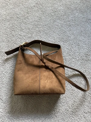 Brun mocka axelremsväska - Snygg handväska i mjuk brun mocka med justerbar axelrem i läder och guldfärgat spänne. Väskan har en enkel, fyrkantig form och stilren design som passar till många outfits. Bra med utrymme inuti och aldrig använd.