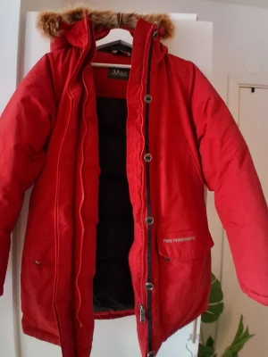 Röd Peak med Dunfyllning  - Snygg röd Peak Performance med huva och avtagbar fuskpäls. Jackan har dragkedja och knappar framtill, flera fickor och varm vaddering. Perfekt för kalla dagar och ger en cool streetstil. 
