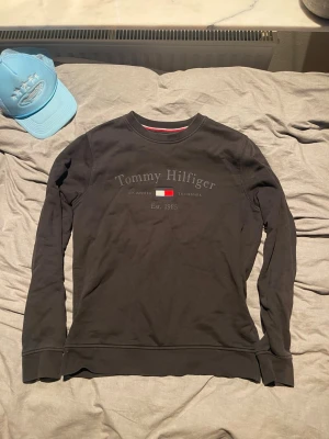 Svart sweatshirt Tommy Hilfiger - Snygg svart sweatshirt från Tommy Hilfiger med tryckt logga och flagga på bröstet. Tröjan har rund halsringning, långa ärmar och ribbade muddar vid ärmslut och nederkant. Perfekt för en avslappnad och stilren look.