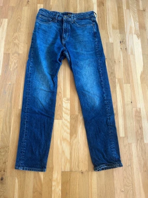 Blå raka jeans från Lee - Storlek 30/32. Klassiska blå jeans från Lee med rak passform och fem fickor. Jeansen har en snygg tvättad look och Lee-logga på knappen och bak i linningen. Tillverkade i slitstark denim, perfekta för en avslappnad stil.