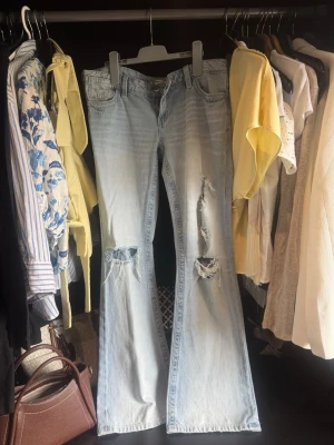 Ljusblå slitna jeans - Säljer ett par ljusblå jeans med flera slitningar och hål på benen. Jeansen har låg midja och klassisk femficksdesign. Perfekta för en avslappnad och trendig look. Materialet är denim i bomull.