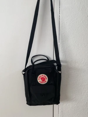 Svart Kånken axelväska mini - Säljer en svart Kånken axelväska mini från Fjällräven. Väskan har justerbar axelrem, två handtag upptill och en liten ficka framtill med knapp. Klassisk rund logga i rött och vitt på framsidan. Tillverkad i slitstarkt material.