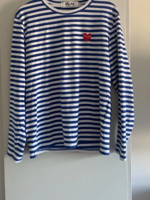 Randig långärmad tröja Comme des Garçons - Säljer en långärmad tröja från Comme des Garçons PLAY med blå och vita ränder. Tröjan har ett rött hjärta med ögon broderat på bröstet och rund halsringning. Perfekt för dig som gillar streetwear och klassisk design. Storleken är L men passar litet. Hör av dig vid frågor eller funderingar.
