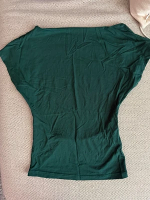 Mörkgrön topp med öppen rygg - Snygg mörkgrön topp i mjukt material med korta ärmar och öppen rygg. Perfekt för dig som vill ha en enkel men ändå unik topp till din outfit. Passformen är normal och tyget känns lätt och skönt mot huden.