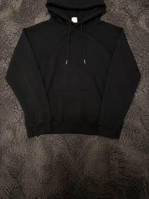 Svart hoodie från H&M Basic - Klassisk svart hoodie från H&M Basic med huva och dragsnören. Hoodien har en stor magficka och ribbade muddar vid ärmslut och nederkant. Perfekt för en avslappnad och stilren look. Tillverkad i mjukt material för extra komfort.