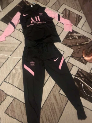 Svart och rosa PSG träningsset Nike - Snyggt svart och rosa träningsset från Nike och PSG. Setet består av en långärmad tröja med rosa ärmar och svarta detaljer samt matchande svarta byxor med rosa inslag. Perfekt för fotbollsträning eller sportiga dagar.