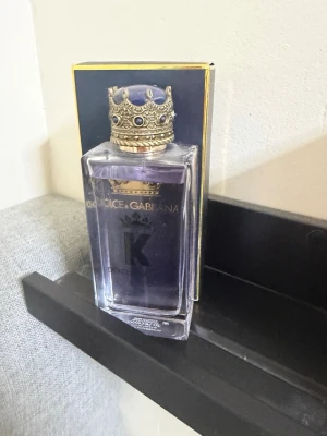Dolce & Gabbana K Eau de Toilette - Snygg parfymflaska från Dolce & Gabbana med lila doftvätska och en unik kronformad kork i guld med blå detaljer. Flaskan är rektangulär och genomskinlig, vilket ger en lyxig känsla. Perfekt för dig som vill ha en elegant och modern doft.
