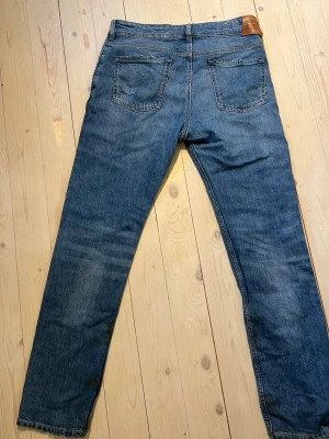 Blå raka jeans med rå kant - Säljer ett par klassiska blå jeans med rak passform och rå, fransig kant vid bensluten.  en läderpatch bak i midjan. Tillverkade i slitstark denim, perfekta för en avslappnad och trendig look. Jensen är använd mindere än 5 gånger jag har slitit dom själv. ag är 172 och jeansen passar bra o h funkar för länge än 172. 