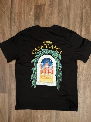Svart t-shirt från Casablanca - Svart t-shirt från Casablanca med färgglad tryckt grafik på både fram- och baksidan. Motivet visar en fönsterliknande illustration med gröna blad och texten 'Avenida Casablanca' i gult. Klassisk passform och rund hals.
