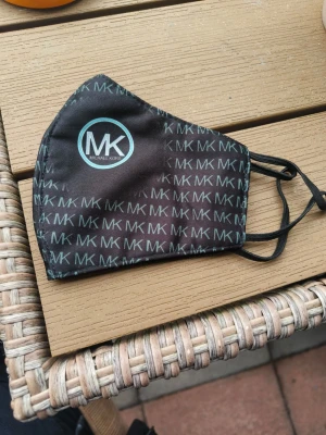 Svart ansiktsmask Michael Kors - Snygg svart ansiktsmask från Michael Kors med MK-logga i ljusblått tryck över hela masken. Masken har elastiska öronband och är tillverkad i ett mjukt tygmaterial. Perfekt accessoar för dig som vill ha stil även när du bär munskydd. OBS knappt använd och tvättad ( svart och grön)