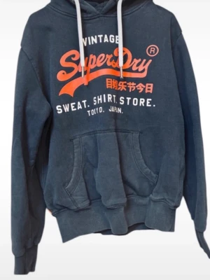Mörkblå Superdry hoodie med tryck - Snygg mörkblå hoodie från Superdry med stort orange och vitt tryck på bröstet. Klassisk känguruficka framtill, vita dragsnören i huvan och ribbade muddar. Perfekt för en avslappnad och trendig stil.