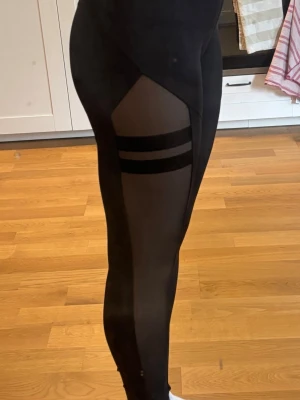 Svarta leggings med mesh och detaljer - Säljer ett par svarta leggings från STRONGER med meshpaneler längs benen och två svarta ränder på låret. Hög midja. Använd ca 5 ggr