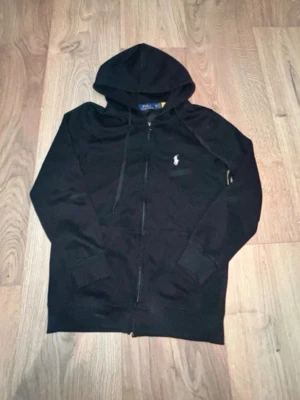 Svart hoodie från Polo Ralph Lauren (M) - Säljer en svart hoodie från Polo Ralph Lauren med dragkedja framtill och klassisk vit broderad logga på bröstet. Hoodien har huva med dragsko och två fickor framtill. Perfekt för en chill och stilren look.