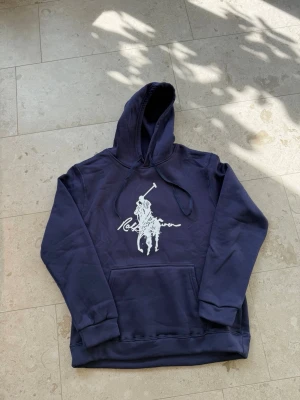 Mörkblå hoodie med stor logga - Snygg mörkblå hoodie från Ralph Lauren med stor vit polospelare och signatur på bröstet. Klassisk känguruficka framtill, ribbade muddar och justerbar huva med snören. Perfekt för en avslappnad och trendig stil.