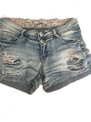 Ljusblå slitna jeansshorts - Säljer ett par ljusblå korta  jeansshorts med slitna detaljer och med low/mid rise. Shortsen har dubbla knappar och dragkedja framtill samt blommigt innerfoder i midjan. Perfekta för sommardagar. Ger lite Y2K vibes och är cirka storlek S. Från Reality jeans denimart.