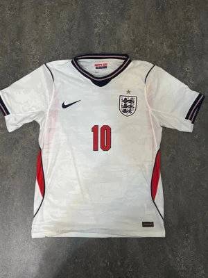 England Home Tröja vm-26 - Som ny England fotbollströja med Bellingham #10  Tröjan är endast använd en gång och är i mycket fint skick, som ny. Inga skador eller fläckar.  Pris: 500 kr – tycker det är ett bra och rimligt pris för tröjan. Priset kan diskuteras vid snabb affär.  Skickas snabbt och packas ordentligt.