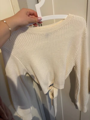 Krämvit stickad tröja med knyt - Säljer en krämvit stickad tröja med rund halsringning och långa ärmar. Tröjan har ett snyggt knytband bak i ryggen och är gjord i ett mjukt, ribbstickat material. Perfekt för dig som gillar en enkel men trendig look.