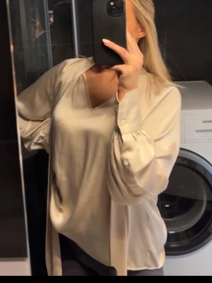 Knytblus från Arket - Snygg beige satinblus med v-ringning och långa, lösa ärmar med breda manschetter. Blusen har en avslappnad passform och ett mjukt, glansigt material som ger en lyxig känsla. Endast använd någon enstaka gång 🩷