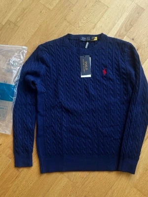 Marinblå kabelstickad Polo Ralph Lauren tröja -  marinblå stickad tröja från Polo Ralph Lauren med rund halsringning och röd broderad logga på bröstet. Tröjan har ribbade muddar vid ärmslut och nederkant. Perfekt för dig som gillar klassisk och stilren design.