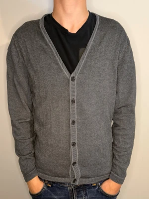 Overshirt - En enkel och stilren grå cardigan med v-ringning och knäppning framtill. Tröjan har långa ärmar, ribbade muddar och är tillverkad i ett mjukt, tunt stickat material. Perfekt att slänga över en t-shirt för en avslappnad look.