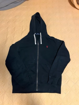 Svart zip hoodie med huva - Säljer en svart hoodie med dragkedja och huva. Hoodien har vita snören och en liten röd logga broderad på bröstet. Perfekt för en avslappnad och stilren look. Materialet känns mjukt och bekvämt.