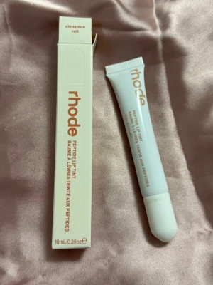 rhode Peptide Lip Tint Cinnamon Roll - Peptide Lip Tint från rhode i färgen Cinnamon Roll. Kommer i en vit tub med guldtext och matchande förpackning. Innehåller 10 ml och har en mjuk applikator. Perfekt för att ge läpparna en hint av färg och återfuktning.