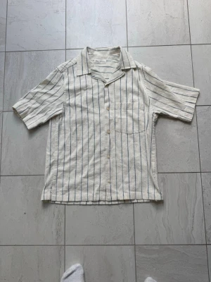  skjorta från Zara - Säljer en ljus, kortärmad skjorta från Zara med tunna blå ränder. Skjortan har klassisk krage, bröstficka och knappar framtill. Tillverkad i ett luftigt linnematerial, perfekt för varma dagar.