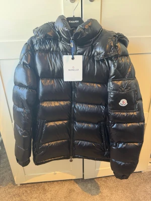 Svart pufferjacka från Moncler - Snygg svart pufferjacka från Moncler med glansig finish och huva. Jackan har två dragkedjeförsedda fickor framtill och en hög krage. Perfekt för kalla vinterdagar och riktigt trendig look.