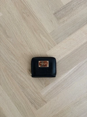 Svart plånbok Michael Kors - Snygg svart plånbok från Michael Kors i läder med guldfärgad dragkedja och metallskylt framtill. Kompakt och fyrkantig modell som är perfekt för kort och kontanter. Enkel och stilren design som passar till det mesta.